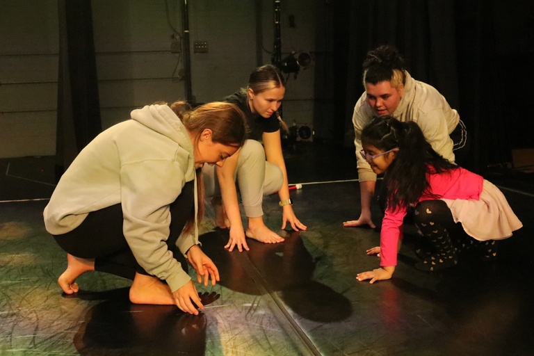 A second grader wearing pink shirt and purple tutu touches the marley floor of Space Place theatre as company members Jemma Hoffman, Gracie Schultzz and research assistant Cami Rezabek teach her choreography from UIDC piece "Winning." 