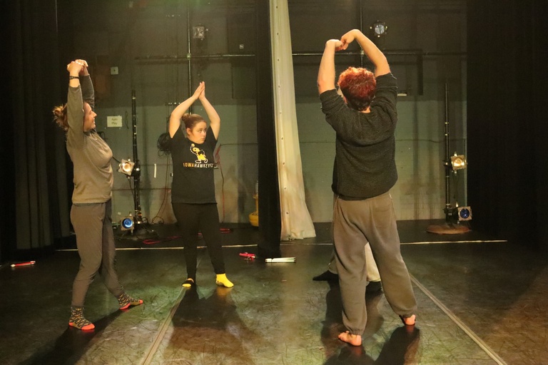 Extended Learning student Brinley leads a group of company dancers as they follow along with her movements. Brinley's, wearing a Hawkeye shirt and gold and black socks, raises her hands high above her head, palms together. Noah and Sophia imitate her movement.