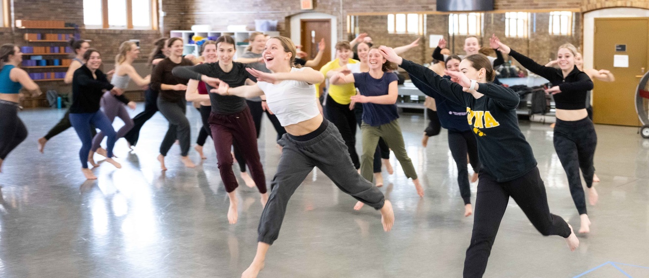 a group of about 20 undergraduate dance students exuberantly moving across the studio floor together during a workshop by Pilobolus dance company
