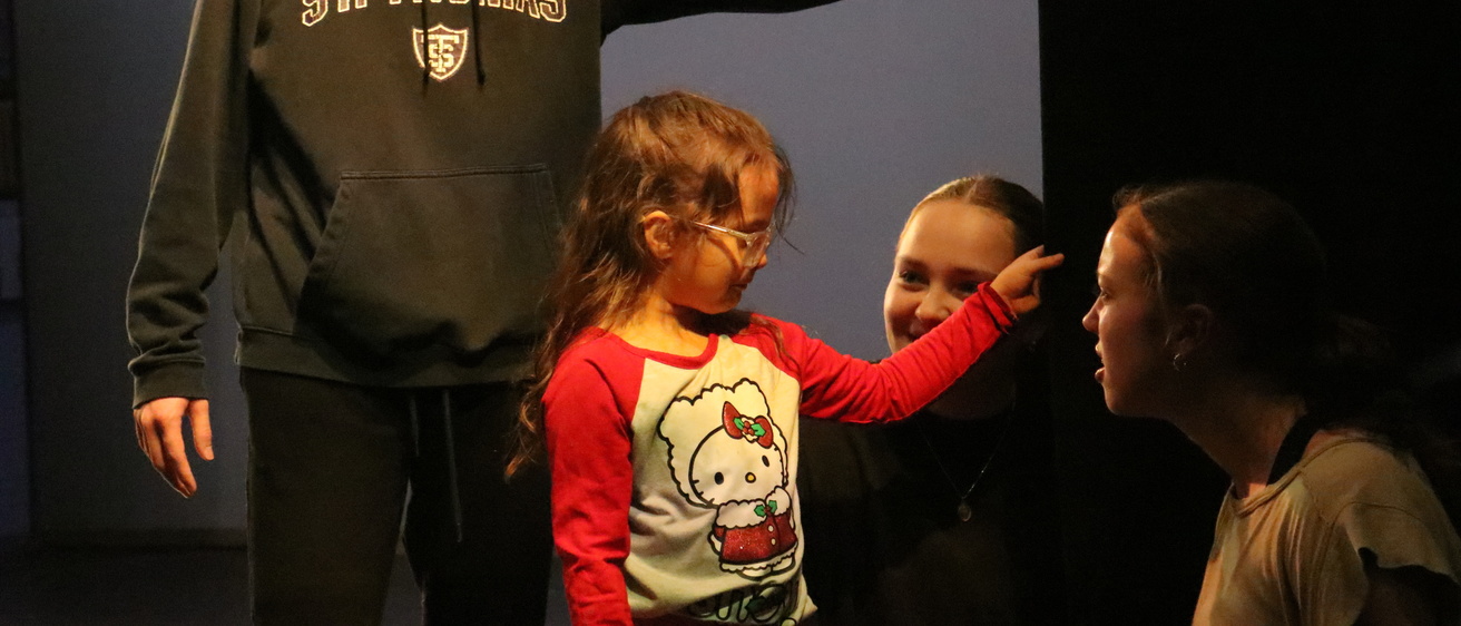 Six-year old EL student wearing a Hello Kitty shirt and pink glasses touches black curtain in Space Place theater as UIDC dancers Jack Delaney (behind in sweatshirt), Ruby Gentzler, and Madison Burkhart (kneeling) listen to her description of the curtain.