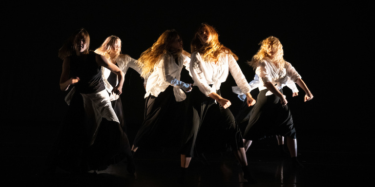 five dancers dressed in black and white, with long blonde and red hair in motion, against a black background and lit dramatically