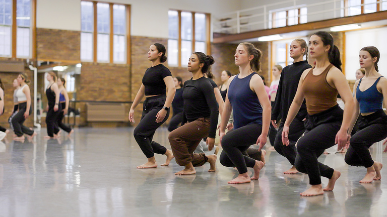 UI Dancers divided into groups for a run through of Martha Graham's Panorama. Group in focus is in a squatting lunge with the left leg forward and arms to the side in a position that might look like a Barbie doll's natural pose.