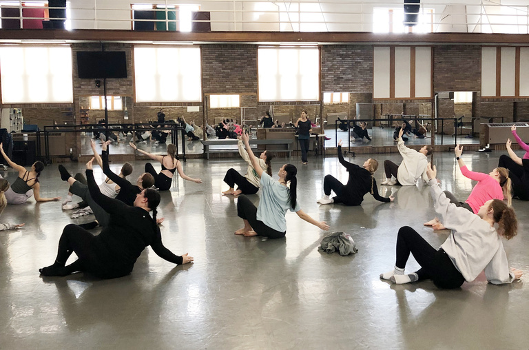 Virginie Mécène leading UI dancers in a workshop on Graham Technique. Dancers are on the floor, balancing on their right hip, feet together, legs bent, and arms outstretched. Left arms are raised above their bodies and right arm extended to the floor.