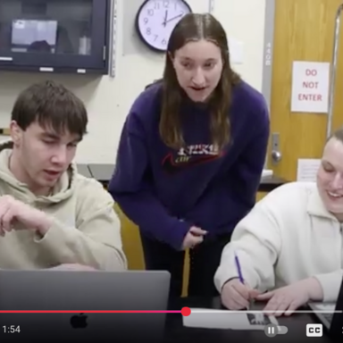 three students in an anatomy lab, looking together at their laptop screens with a skeleton model and chalkboard in the background