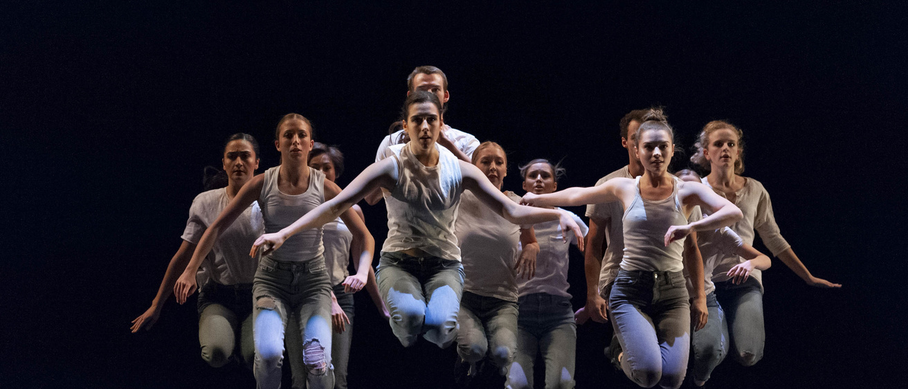 a group of dancers in denim and t-shirts suspended in a jump in unison against a black background