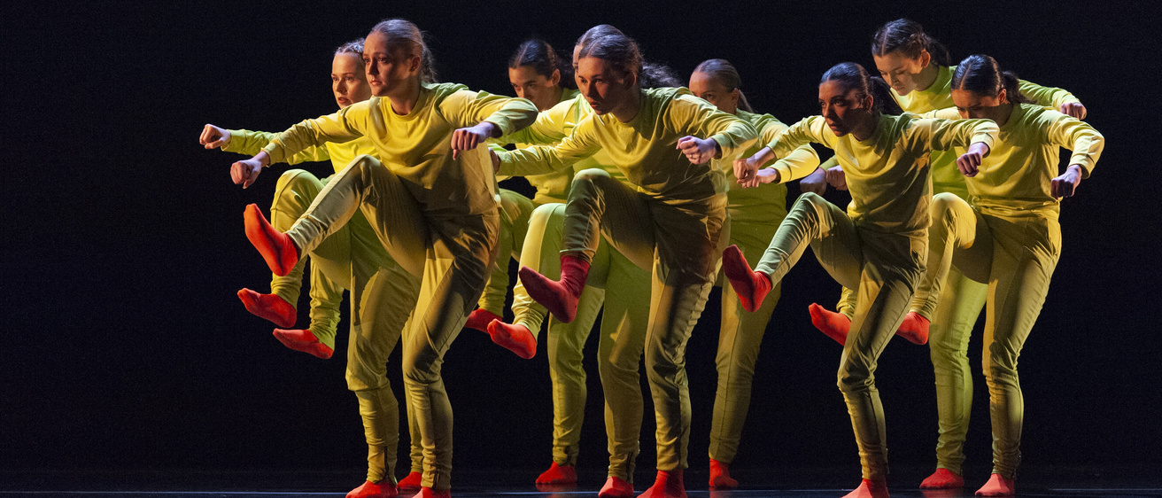 A group of dancers on a dark stage wearing yellow jumpsuits and bright orange socks, with the stage lit in blue shades