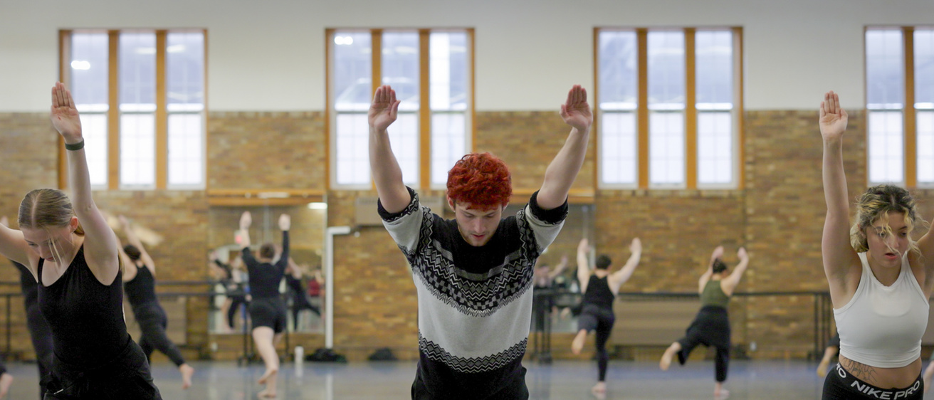 UI Student Dancers in large circle, facing outward, practicing Martha Graham's Panorama. There are three students in the foreground with arms stretched forward and left leg kicked back so they are balancing on their right. Left is blonde dancer in all black. Middle is a male dancer with bright red hair, black and white sweater over black pants. And to the right is curly blonde and dark hair, white cropped dance shirt and black pants. 