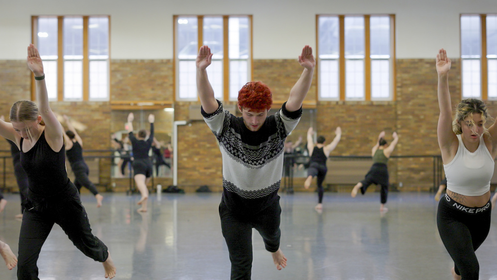 UI Student Dancers in large circle, facing outward, practicing Martha Graham's Panorama. There are three students in the foreground with arms stretched forward and left leg kicked back so they are balancing on their right. Left is blonde dancer in all black. Middle is a male dancer with bright red hair, black and white sweater over black pants. And to the right is curly blonde and dark hair, white cropped dance shirt and black pants. 