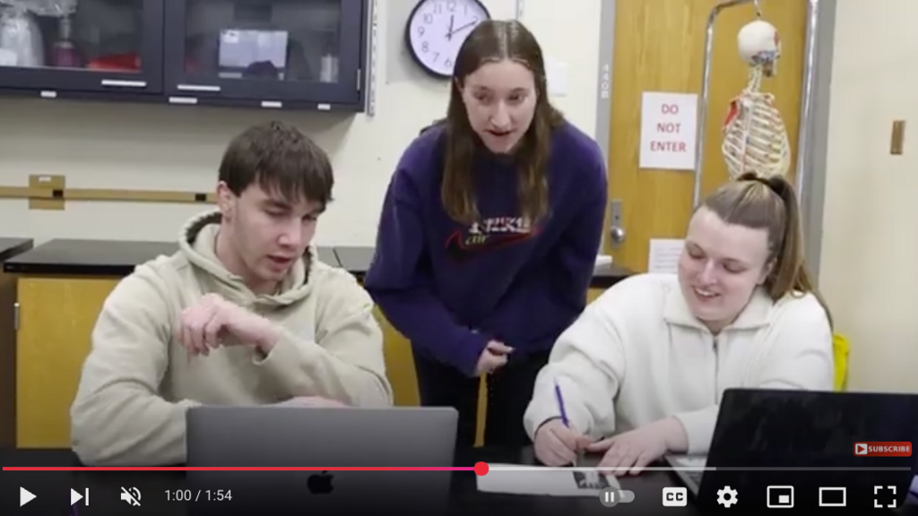 three students in an anatomy lab, looking together at their laptop screens with a skeleton model and chalkboard in the background